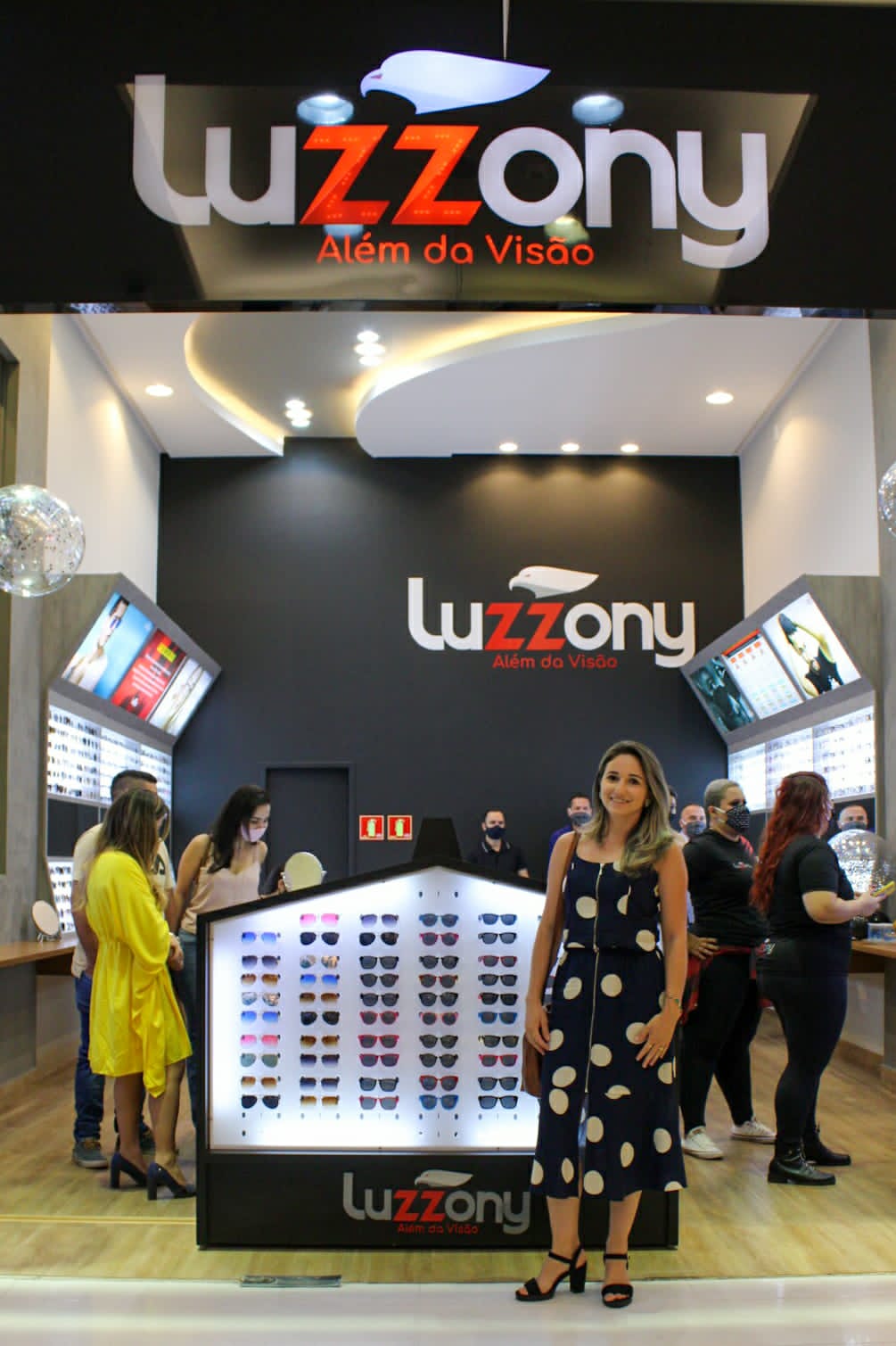 Fachada da loja Óticas Luzzony no Pátio Central com letreiro iluminado, forro curvo e expositor central em formato de casa.